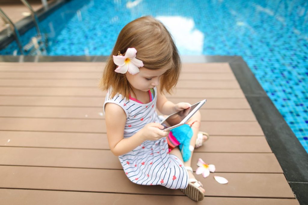 concept of new technologies travel, recreation or holiday family time on sunny day. kid girl uses tablet sitting close to blue swimming pool wearing flower and hold toy